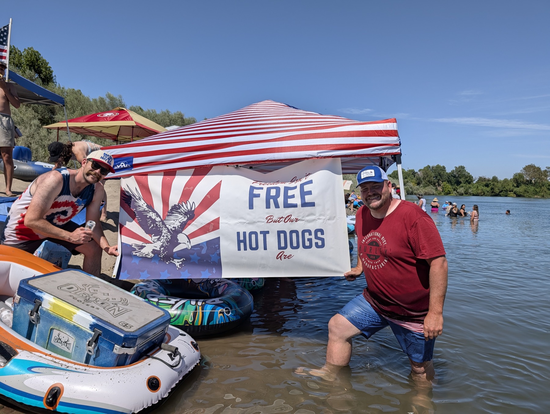 Two men holding the Freedom Isn't Free But Our Hot Dogs Are banner in front of the stars-and-stripes canopy on the Sacramento River
